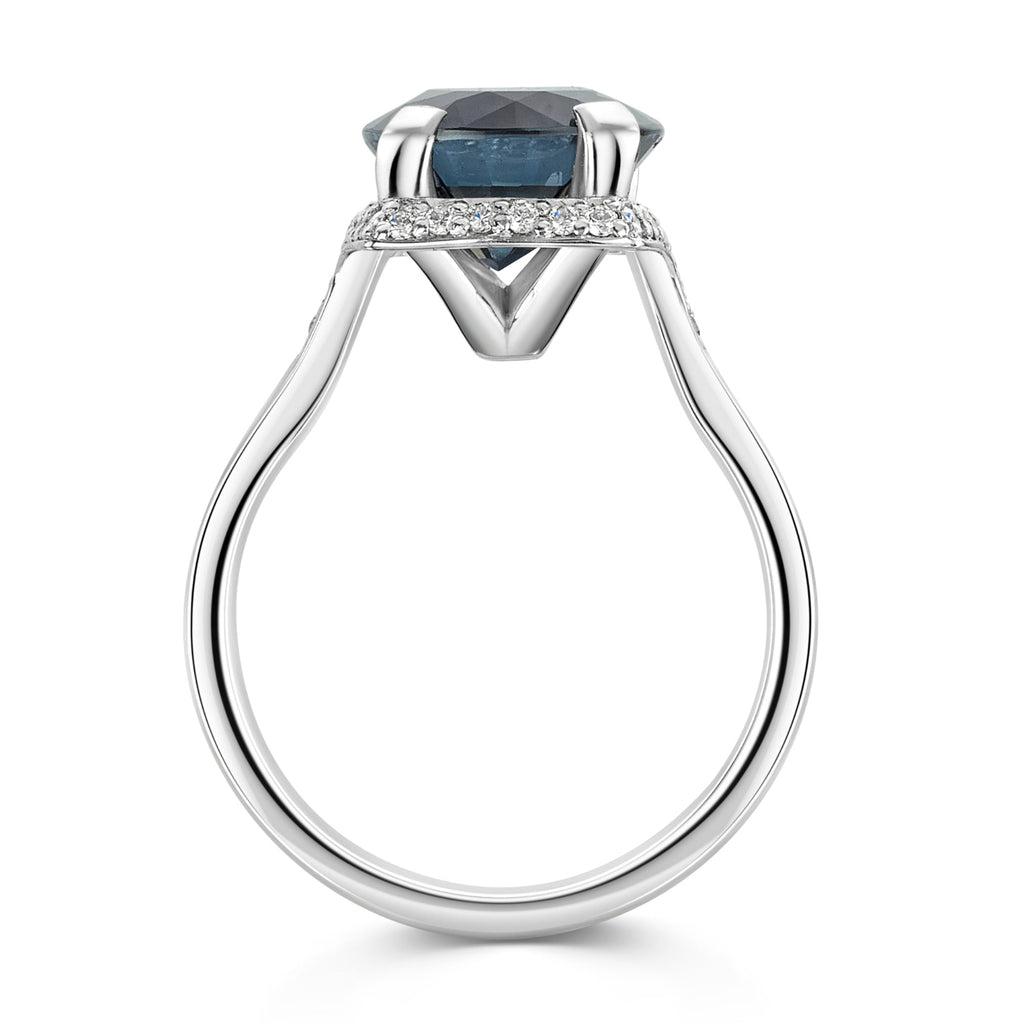Parti sapphire in blue and grey tones, surrounded by a halo of round brilliant cur diamonds set in a platinum band on a white background - side