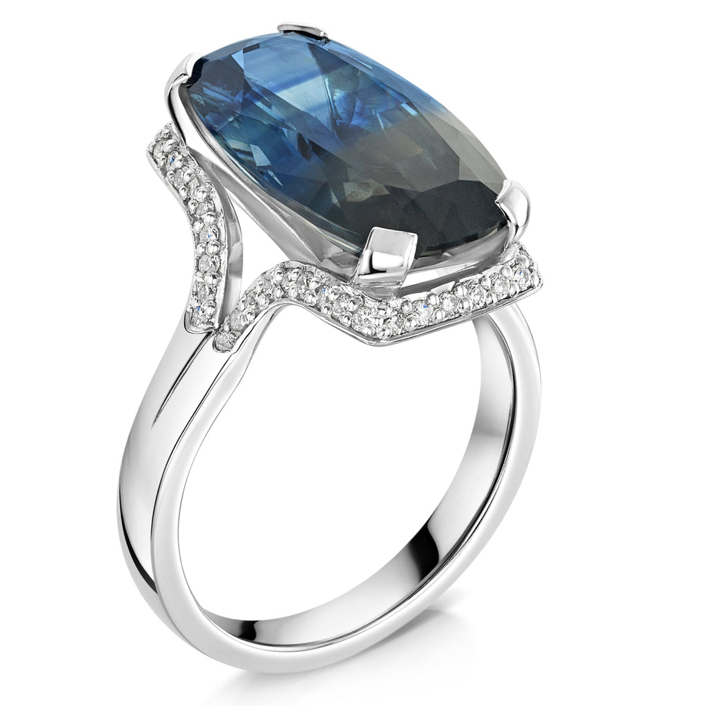 Parti sapphire in blue and grey tones, surrounded by a halo of round brilliant cur diamonds set in a platinum band on a white background - side angle