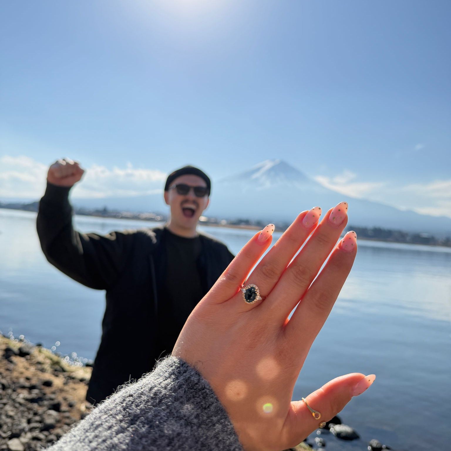 A close-up of a hand wearing a silver ring with a dark oval stone, set against a scenic background of a lake, distant mountain, and a smiling man in the background under a bright sun.