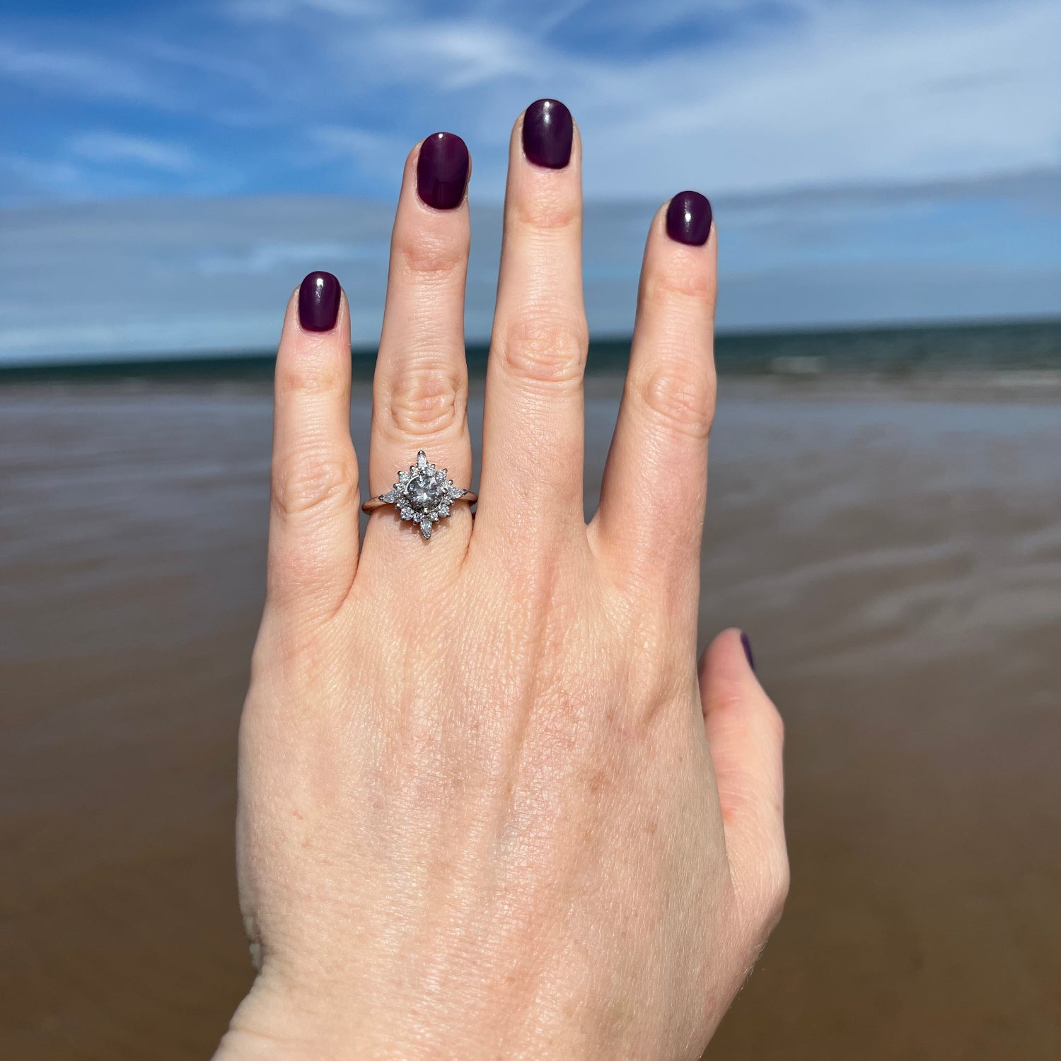 A close-up of a hand with dark purple nail polish wearing a silver-toned ring with a starburst cluster of clear stones, set against a blurred background of blue sky and brown wall.