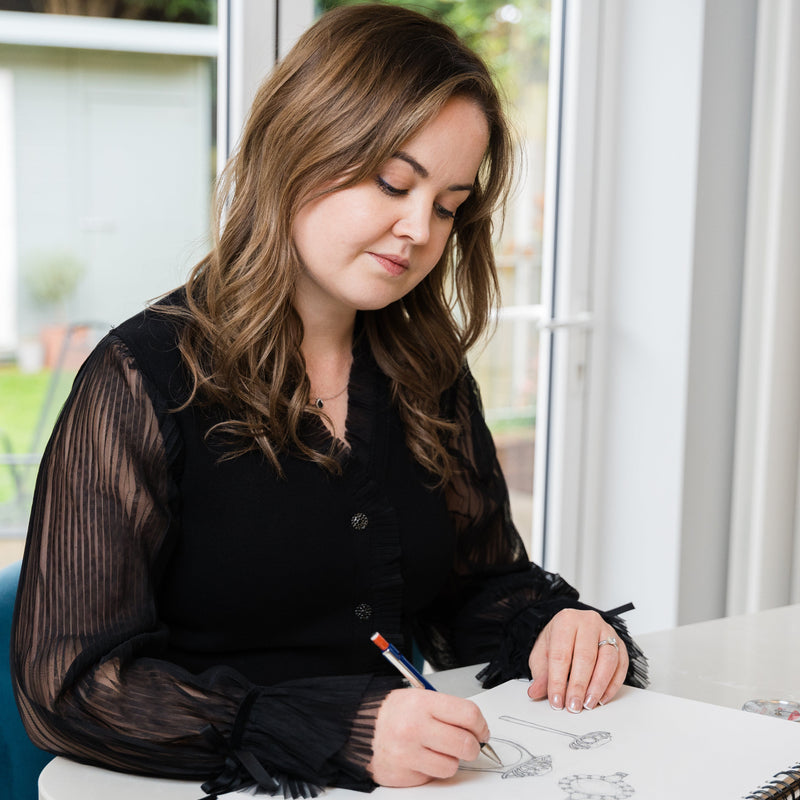 Woman sitting at a desk with a sketchbook and pencil drawing a bespoke engagement ring