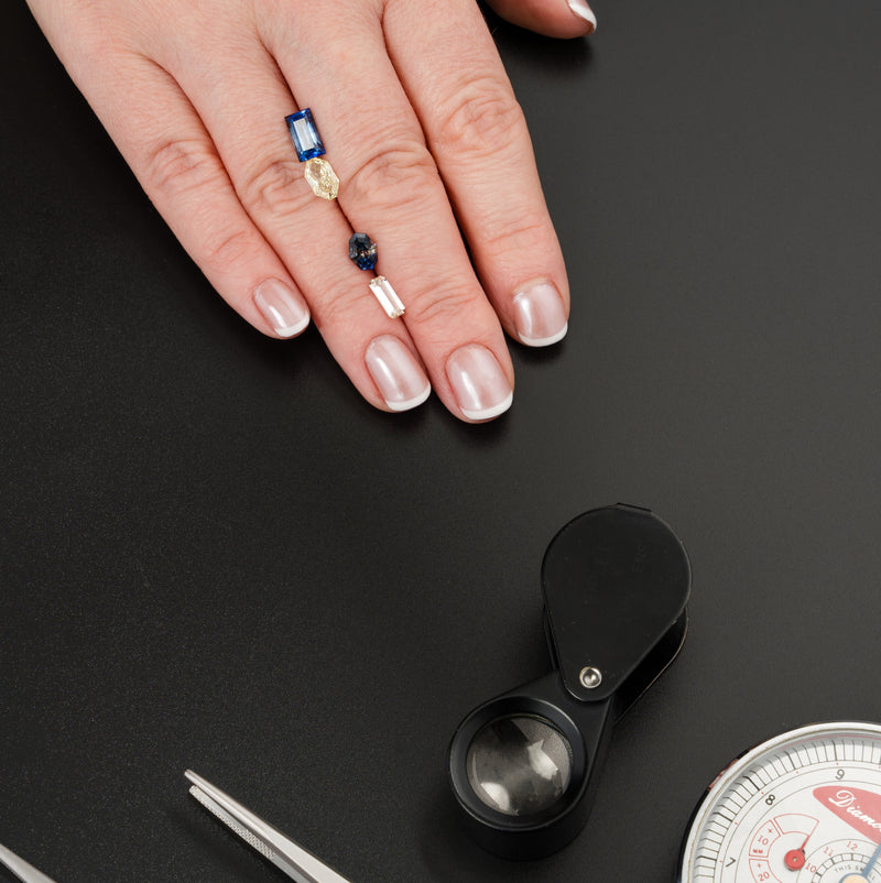 Hand with rings on a black surface with jewelry tools and a pocket scale.