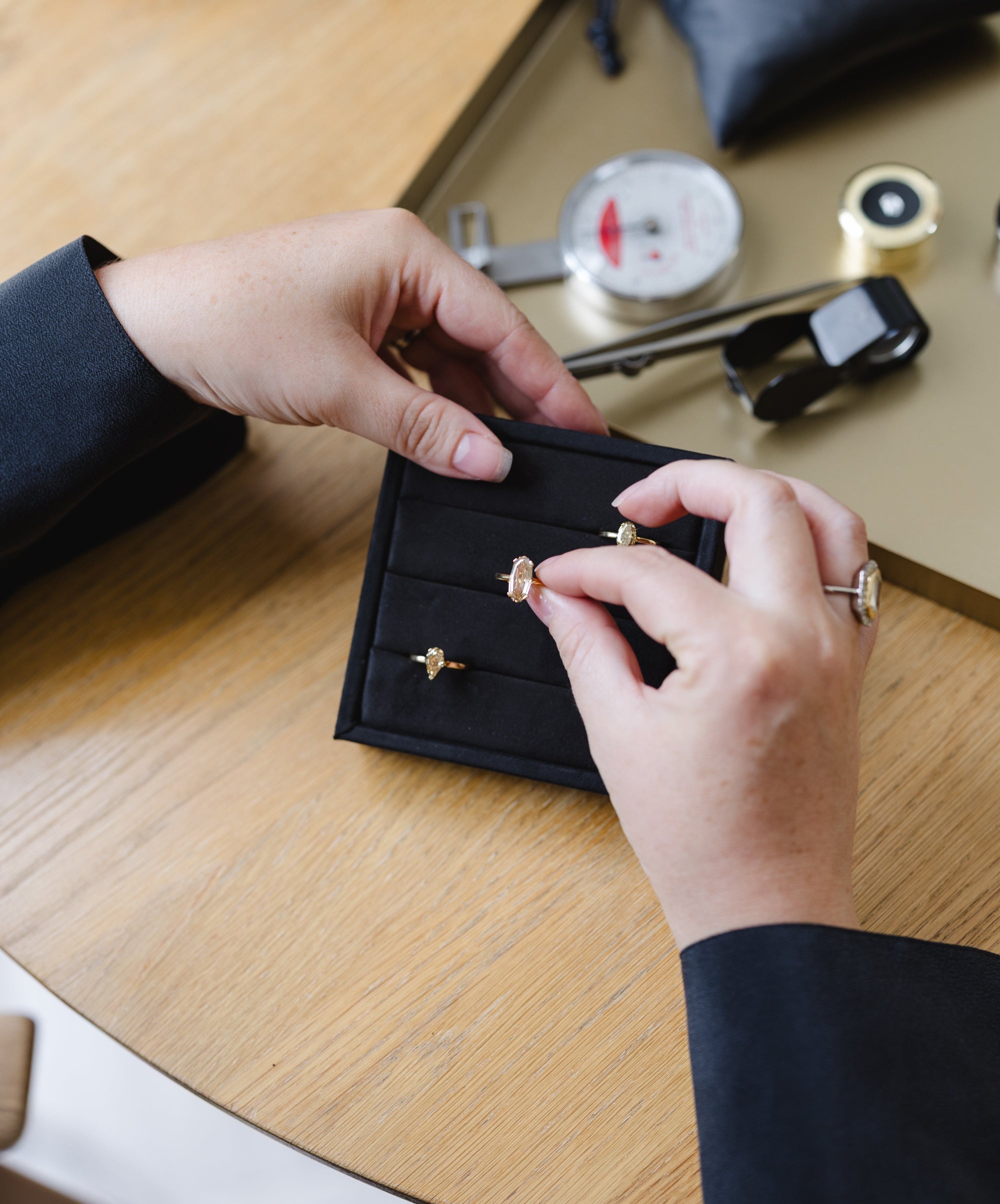 A close-up of a person's hands holding an open black jewelry display card with two small gold rings, set on a light wood table with watch parts and accessories in the background.