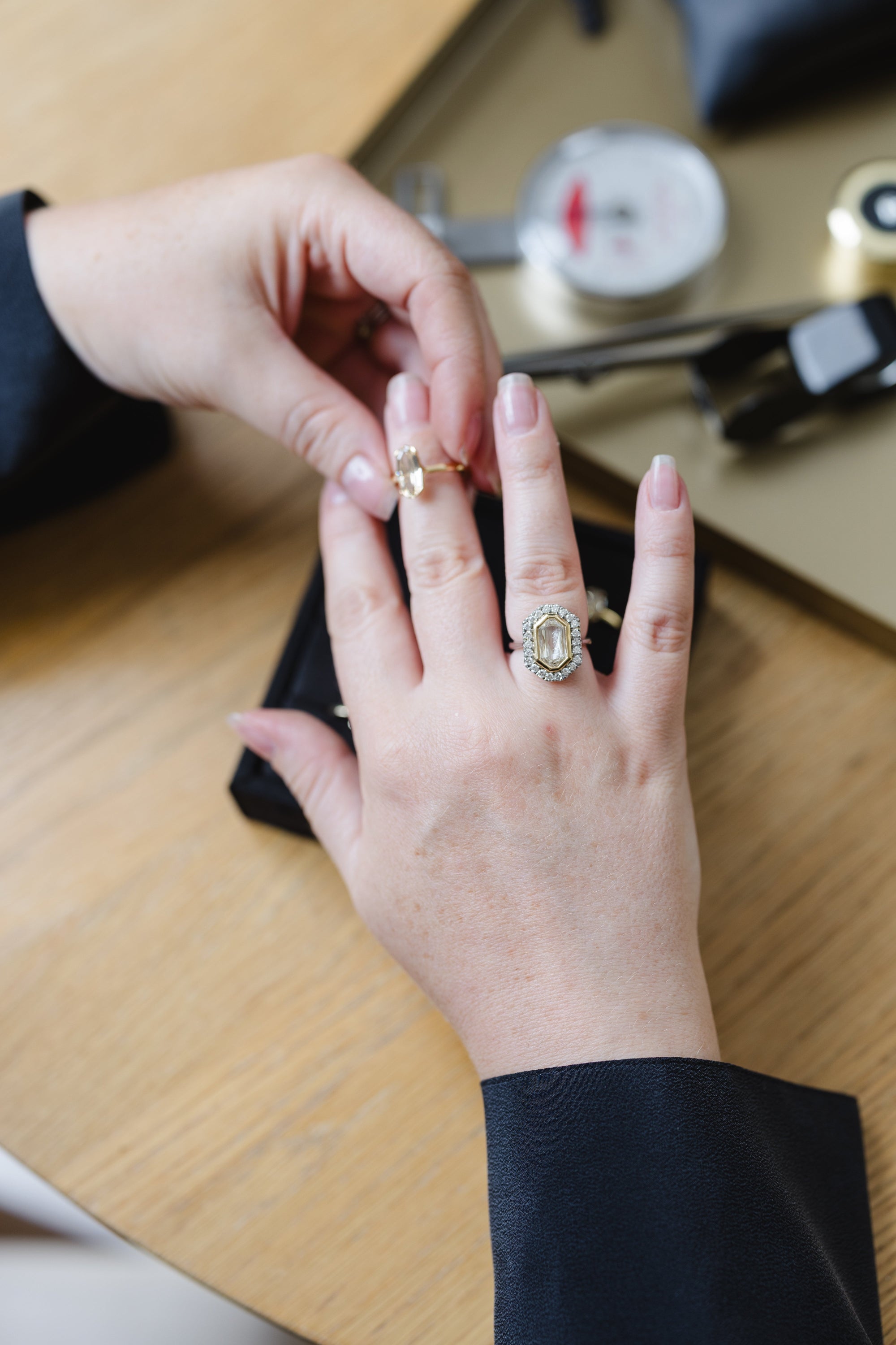 A close-up lifestyle shot of a person's hands wearing two gold rings on their fingers, with one ring featuring a large oval-cut clear gemstone surrounded by a halo of small clear stones. The background shows a wooden table and jewelry tools.