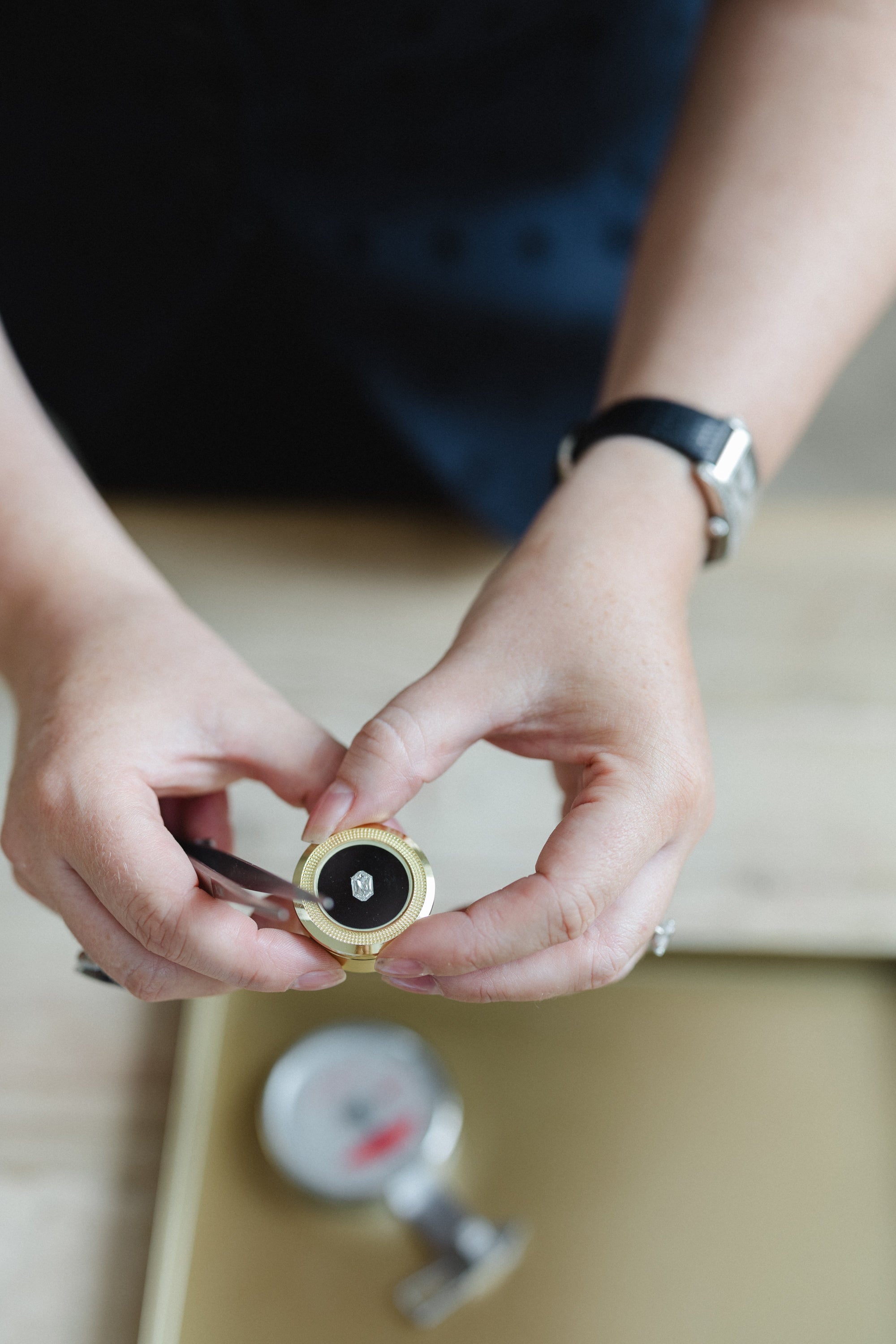 A close-up of a person's hands holding a small round gold-toned pot with a loose hexagonal diamond, against a softly blurred background with a watch on their wrist.