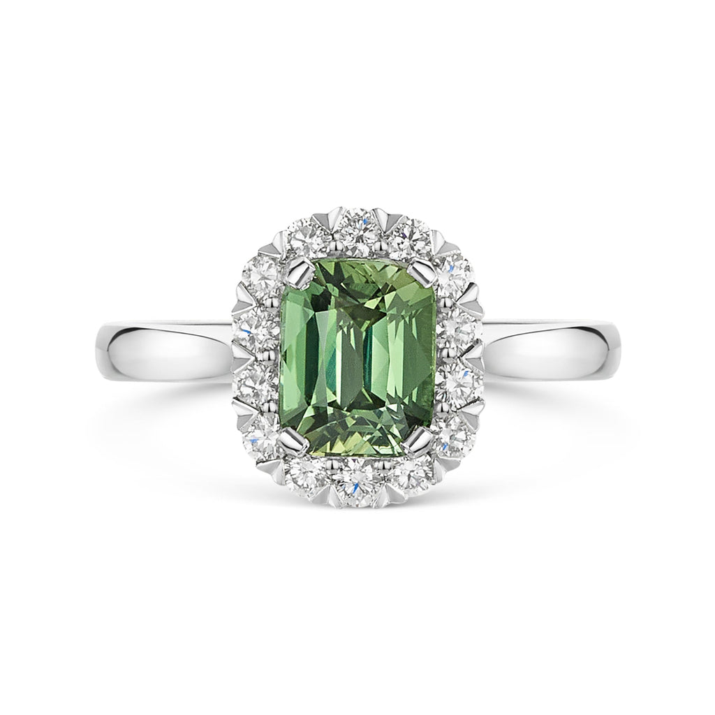 A silver ring featuring a cushion cut green sapphire surrounded by a halo of small white diamonds, set on a polished platinum band against a white background.