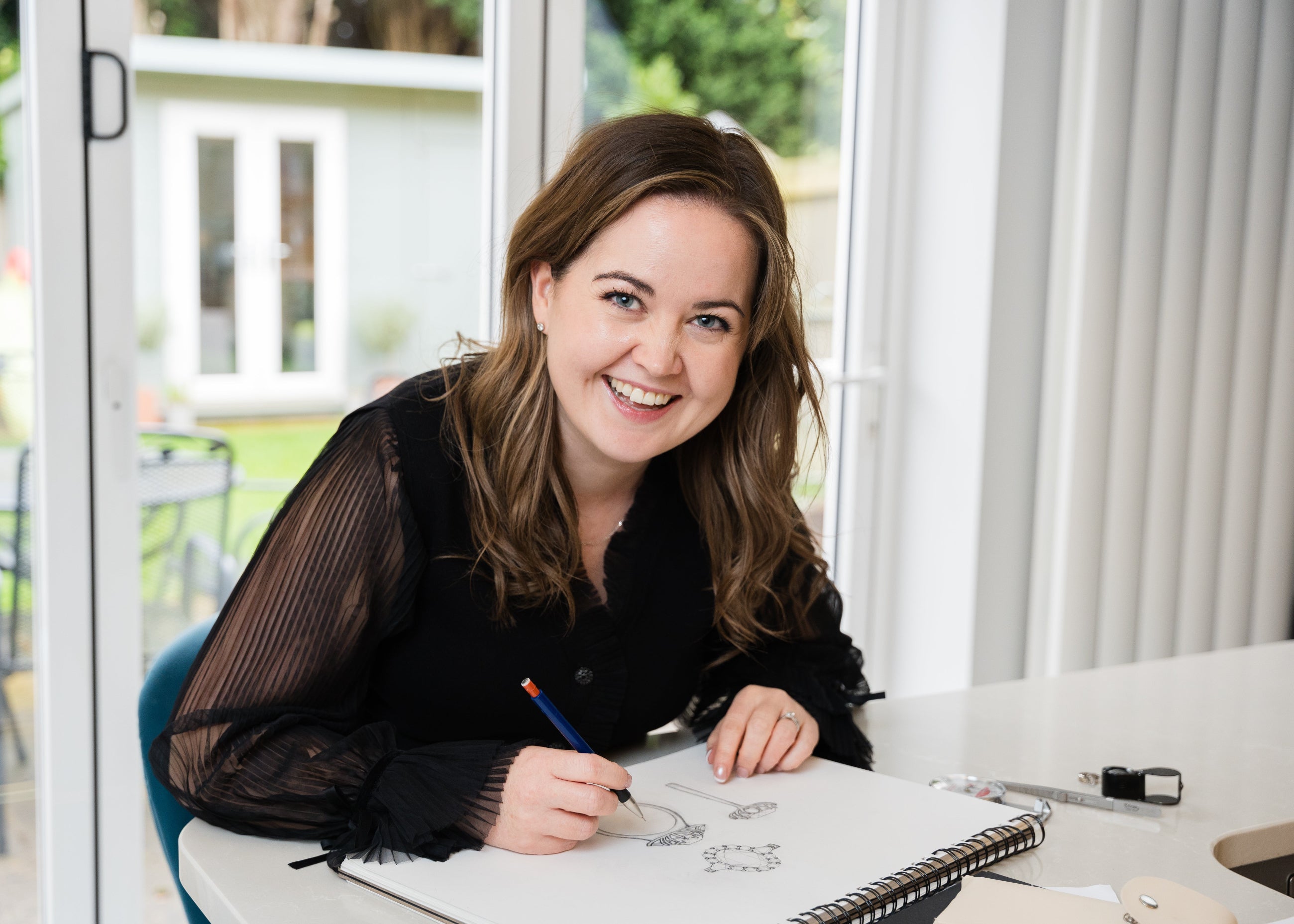 Woman sitting at a desk with a sketchbook and pen, smiling.
