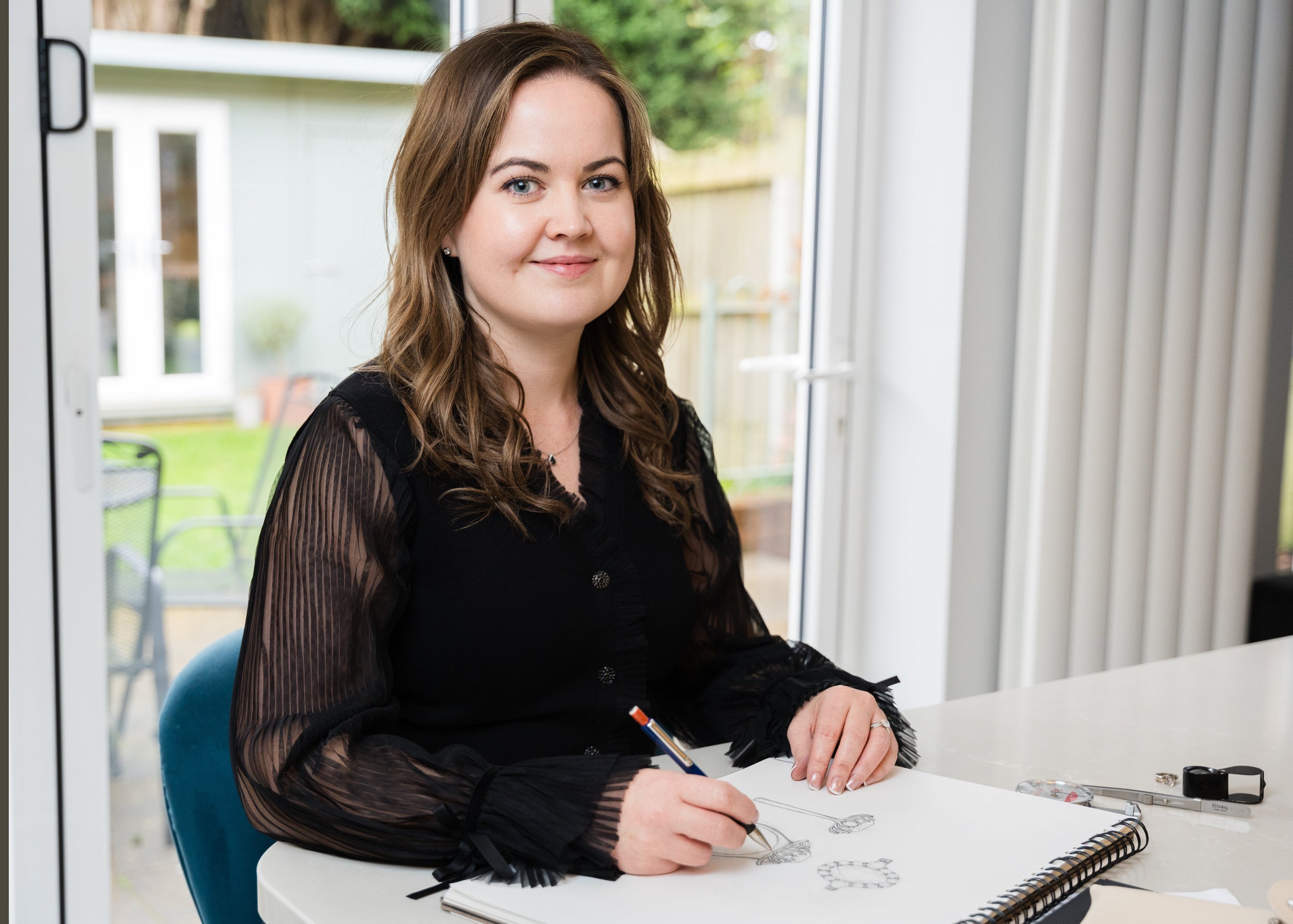 Poppy Elder sitting at a desk drawing in a notebook with a garden view outside.