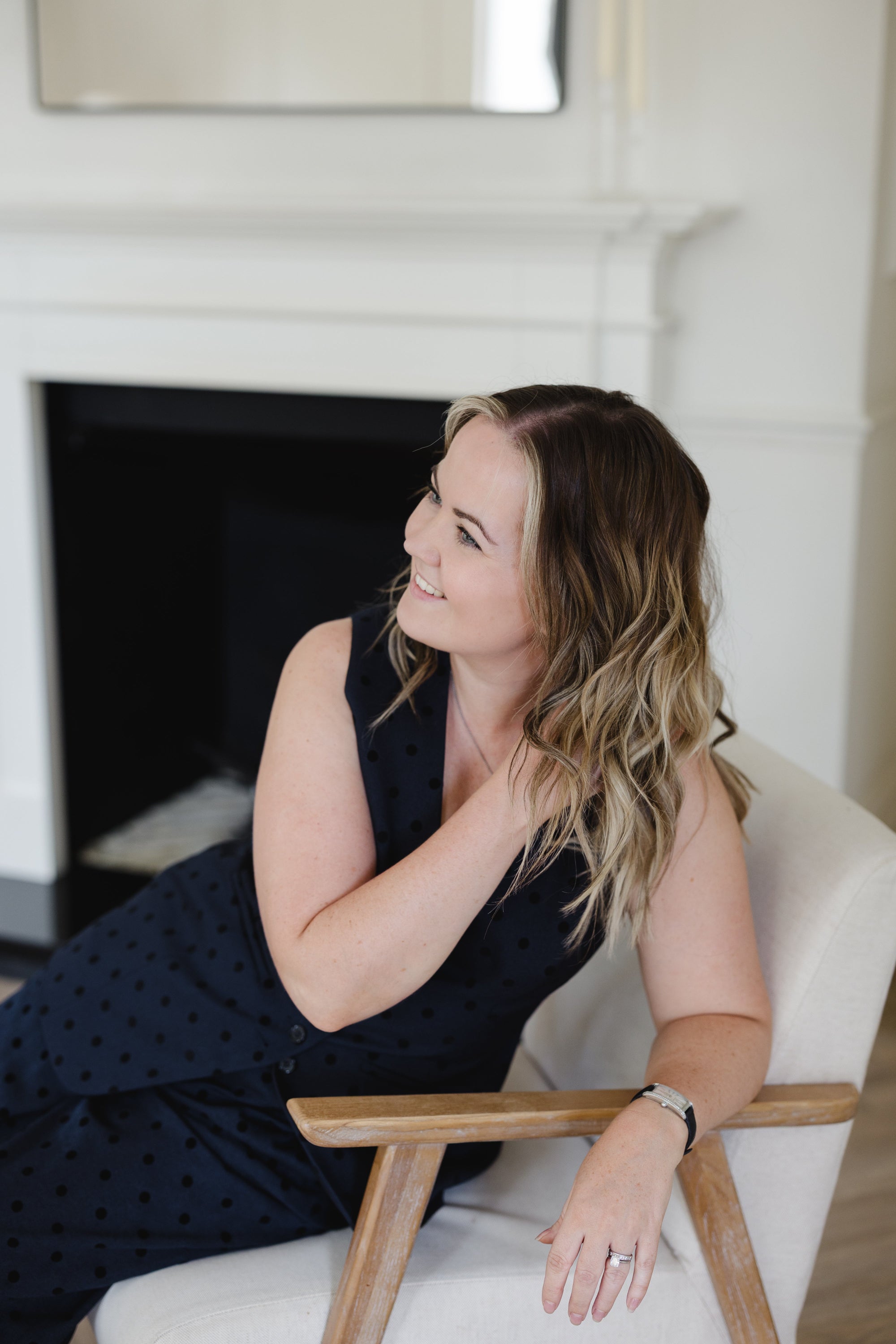 A woman with blonde-highlighted hair wearing a navy blue sleeveless polka-dot dress, sitting in a white armchair with wooden arms in front of a white fireplace.