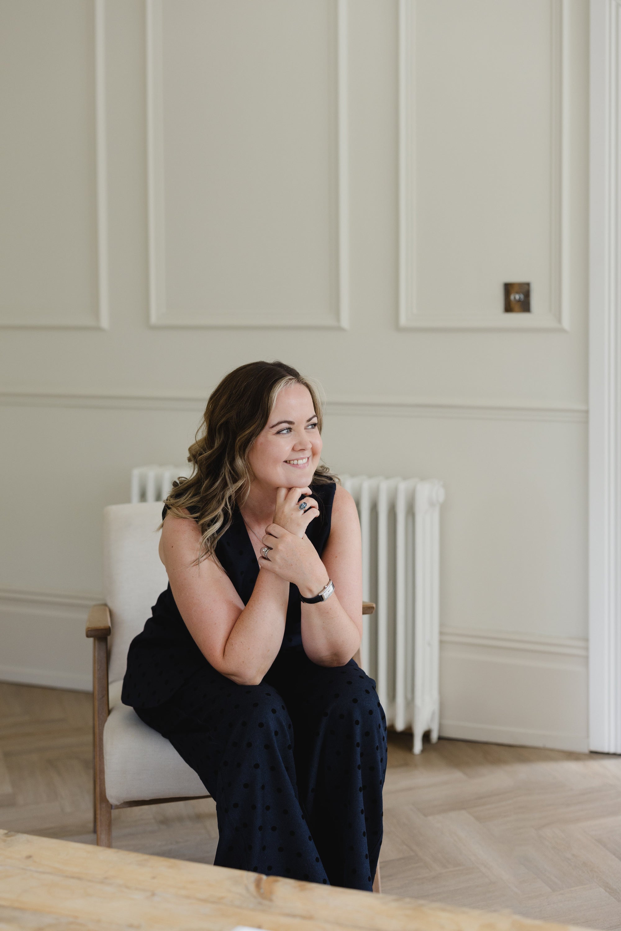 A woman with wavy brown hair sits in a white armchair wearing a black sleeveless top and matching black polka-dot trousers, posing in a bright room with white paneled walls and light wood flooring.