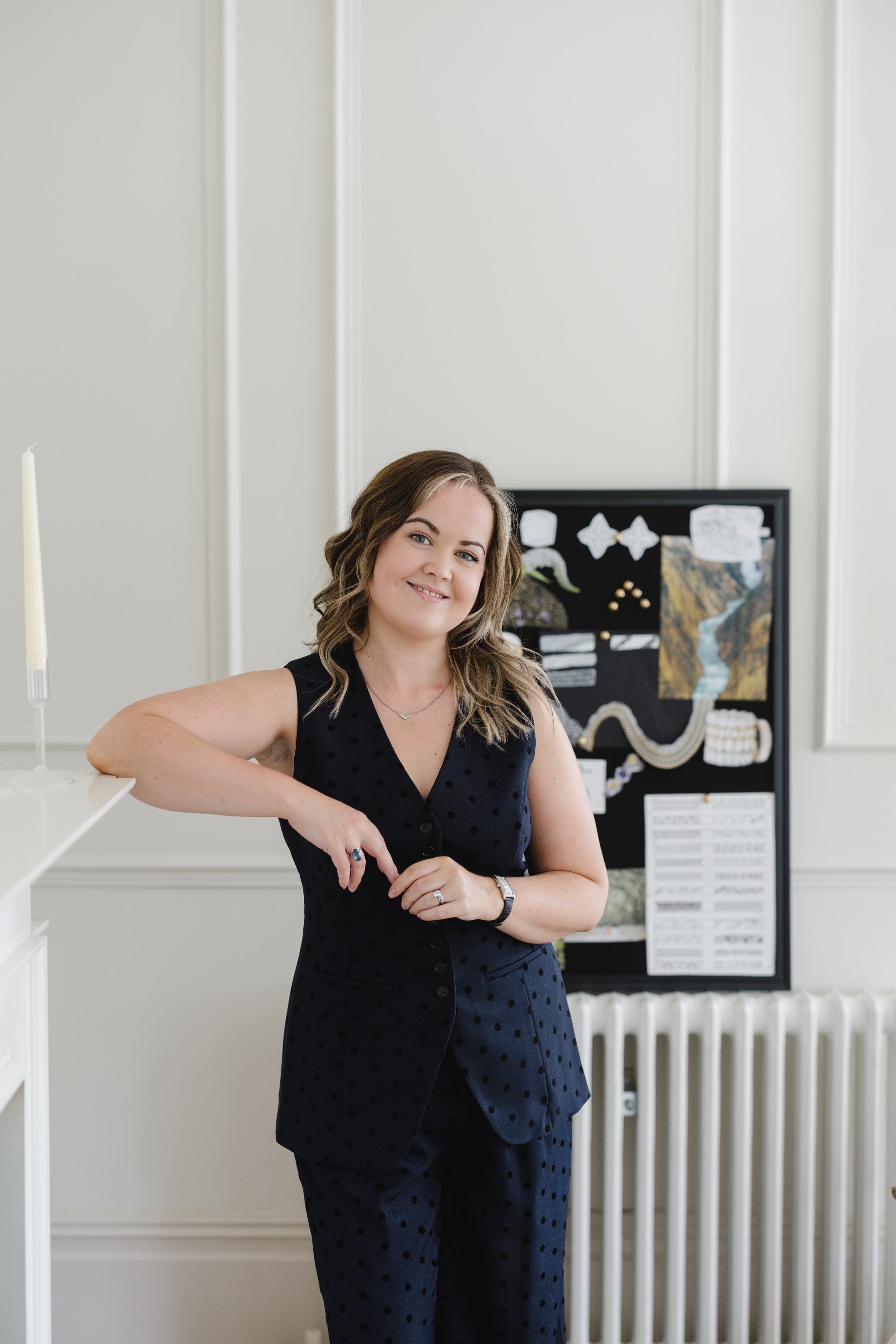 A woman - Poppy Elder - with wavy brown hair stands indoors against a white paneled wall, wearing a dark navy sleeveless polka-dot vest and matching pants. A framed art print hangs on the wall behind her, and a white radiator is visible below.