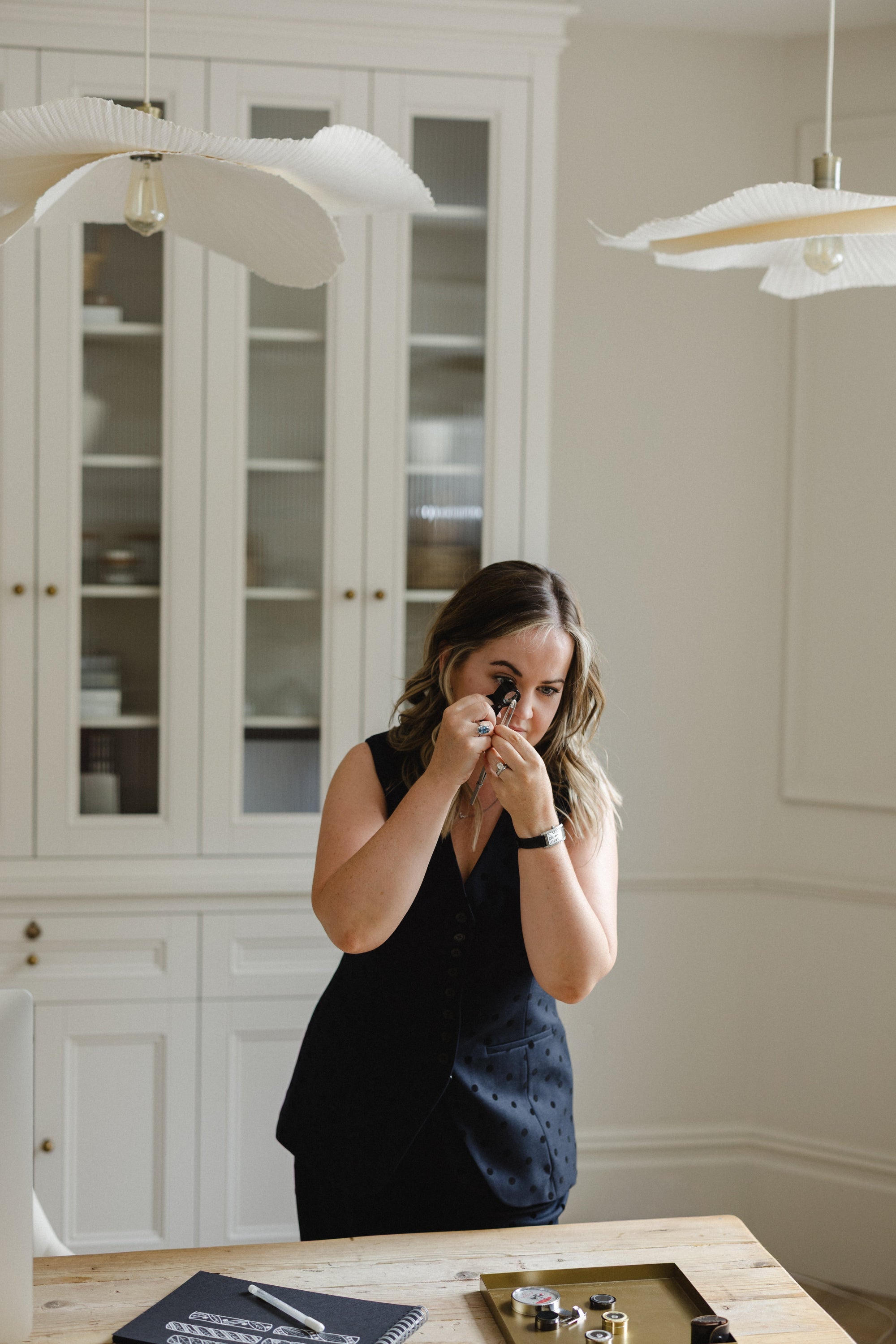 Poppy Elder - inspecting a loose diamond,  in a dark navy sleeveless top stands at a wooden table in a bright, modern room, holding a small magnifying glass. White paneled cabinets and hanging pendant lights are visible in the background.