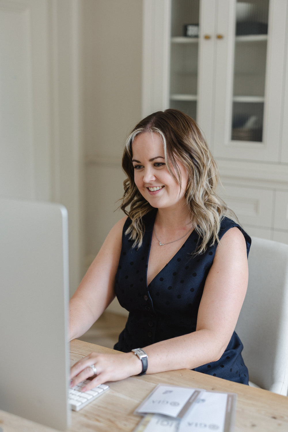 A woman with blonde-highlighted hair sits at a wooden desk using a laptop in a bright, modern home office. She is wearing a dark navy sleeveless top with small white polka dots.