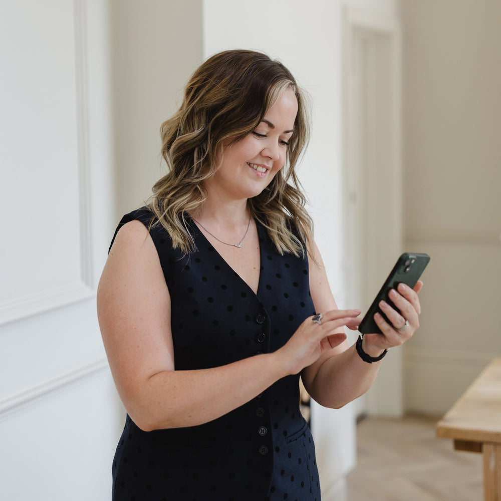 A woman with wavy brown hair stands indoors, smiling at her phone while wearing a sleeveless dark navy polka-dot top. The background shows a bright, modern room with white walls and light wood flooring.