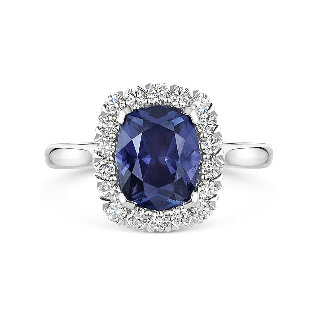 A silver-tone halo ring featuring a large, cushion-cut deep blue purple colour change sapphire center stone surrounded by a halo of small white diamonds, set against a plain white background.