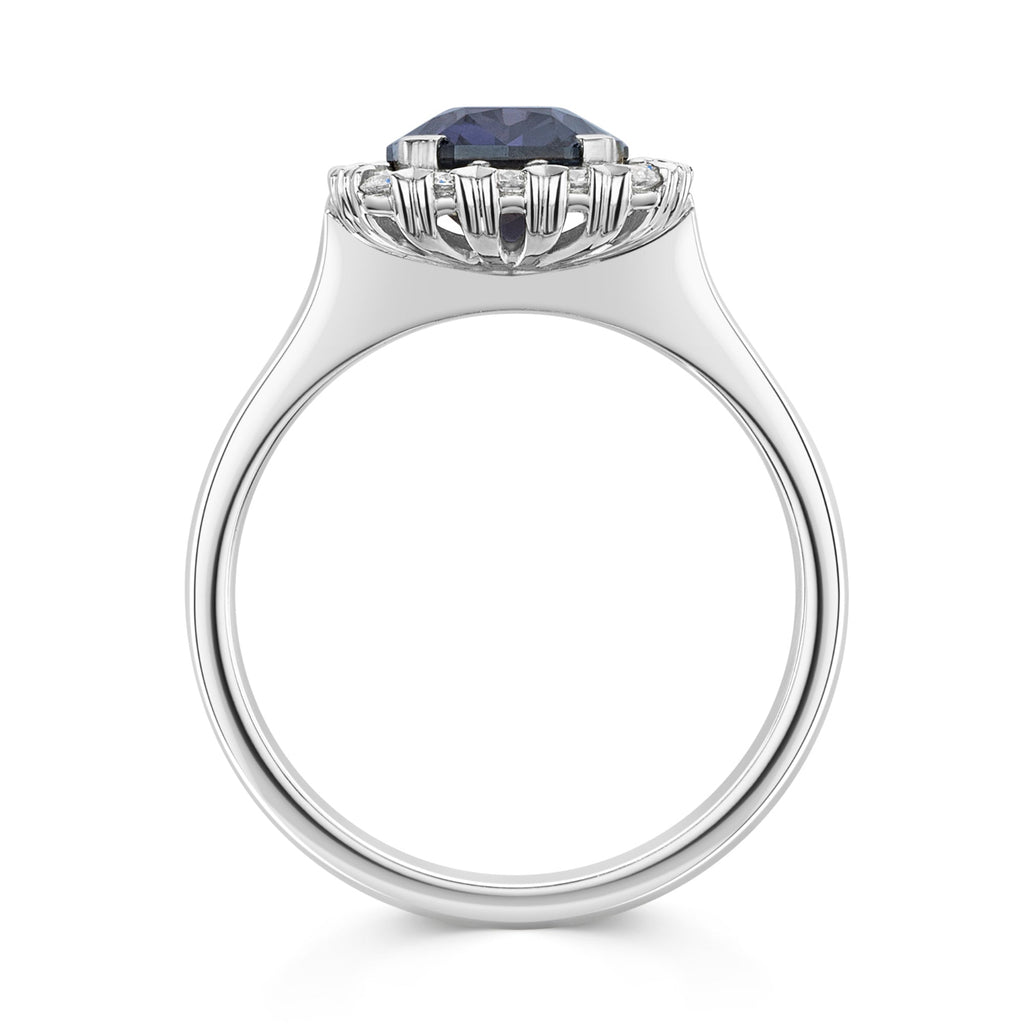A silver-tone halo ring featuring a large, cushion-cut deep blue purple colour change sapphire center stone surrounded by a halo of small white diamonds, set against a plain white background - side view