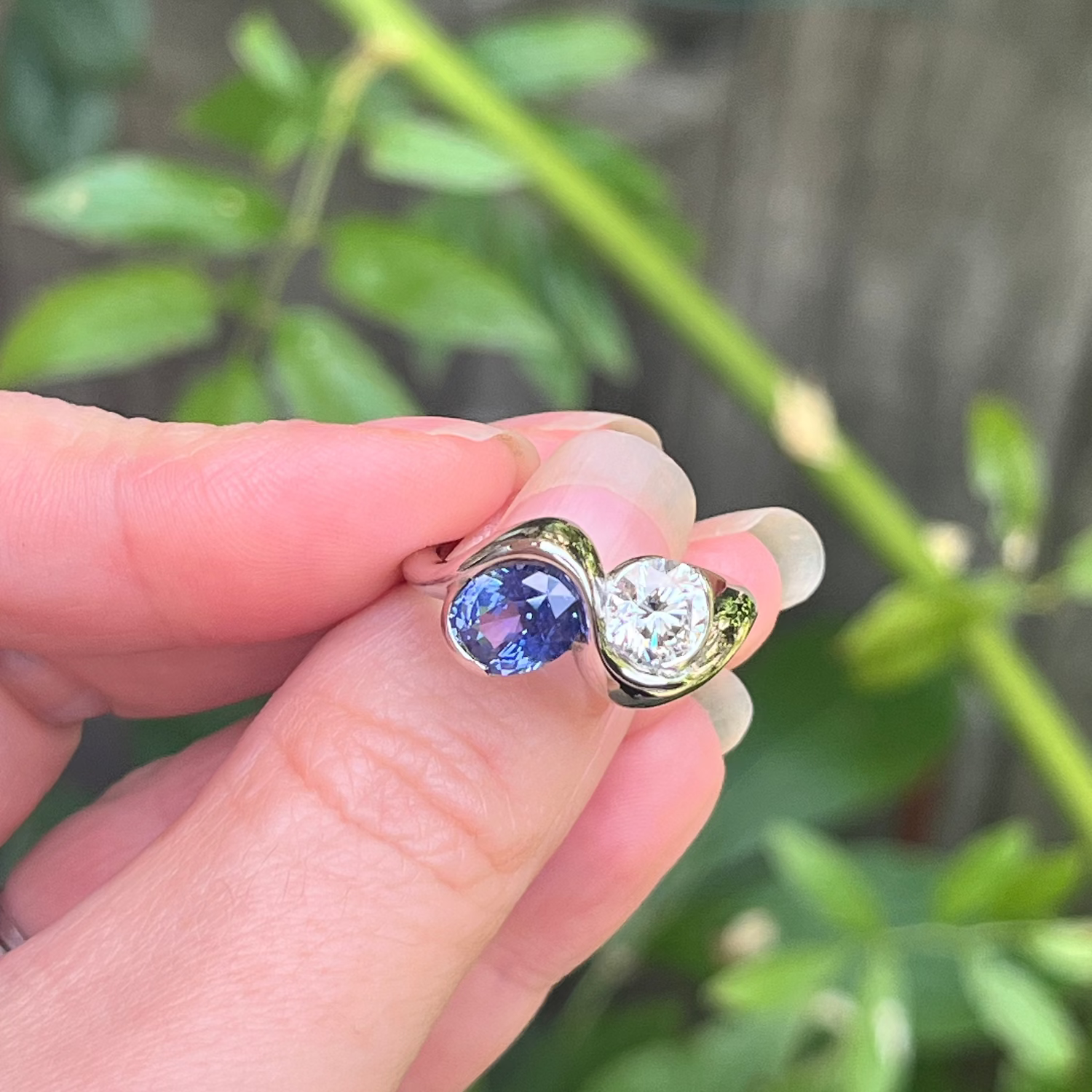 A hand holding a gold ring with a blue oval gemstone and a clear round gemstone, set against a blurred background of green leaves and a wooden fence.