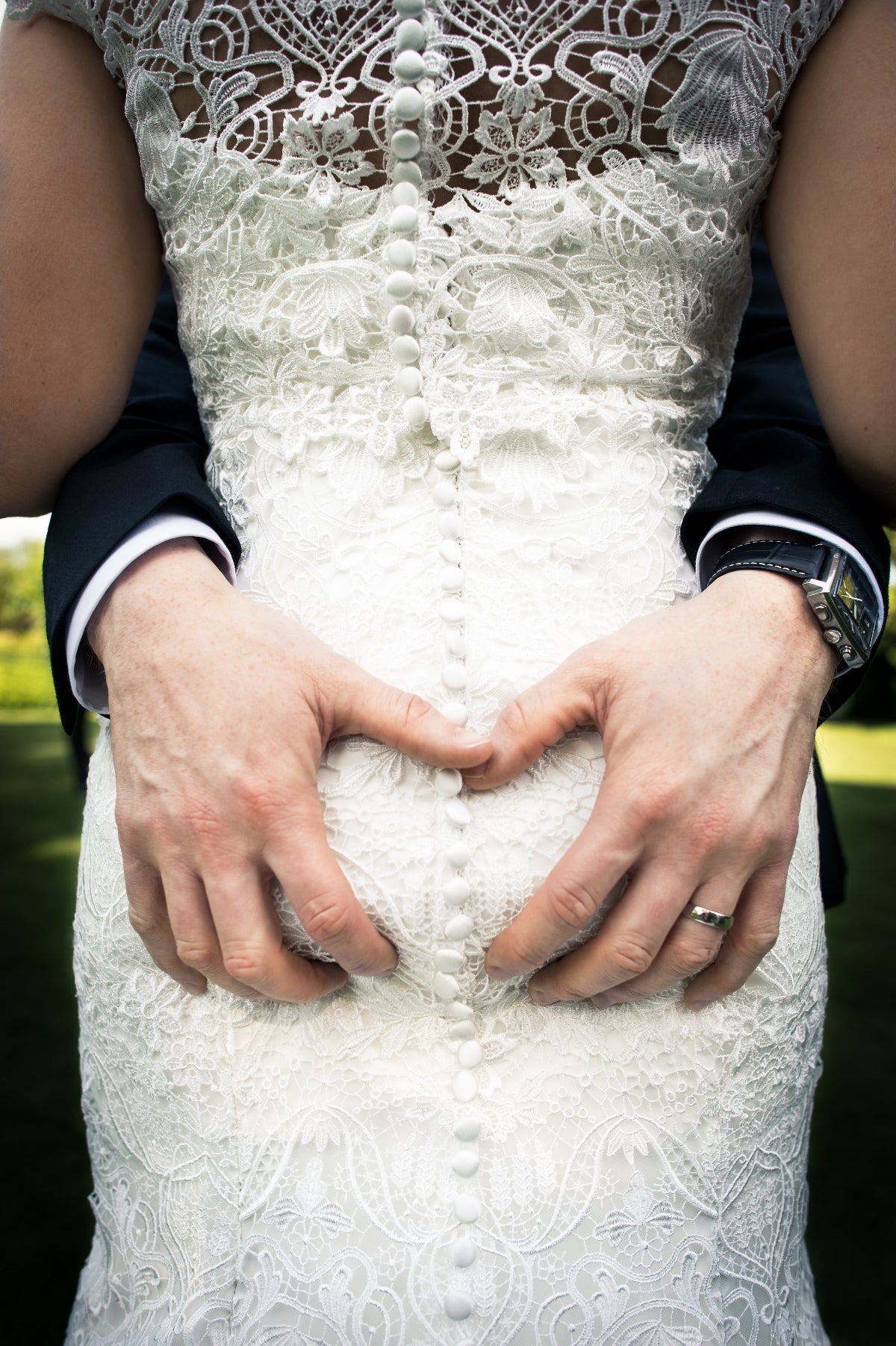 Close-up of hands making a heart shape over a white lace wedding dress.